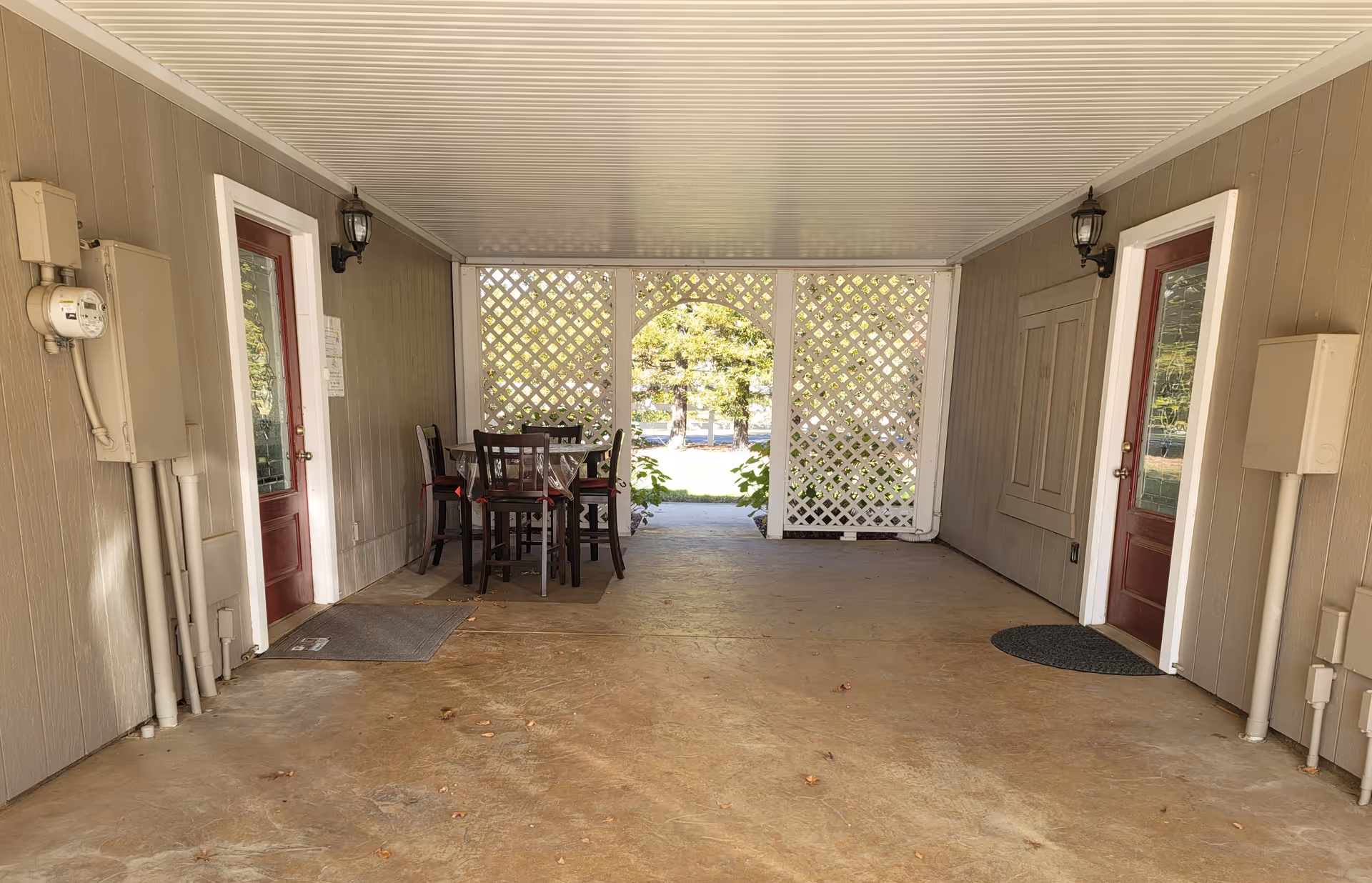 Covered outdoor patio area with two red doors on either side, a small table with four chairs in the corner, and white lattice panels at the far end allowing a view of trees and greenery outside.