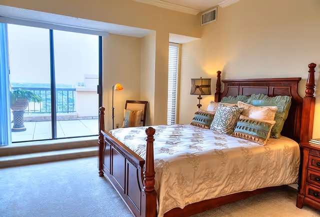 A well-lit bedroom featuring a wooden bed with an ornate headboard and footboard, decorated with multiple pillows and a patterned bedspread. Next to the bed is a wooden nightstand with a lamp. The room has beige walls and carpeted floor. There is a sliding glass door leading to a balcony with a potted plant and a scenic outdoor view.