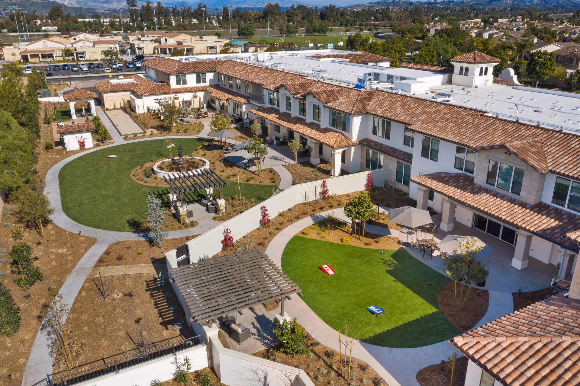 Aerial view of Oakmont of Camarillo senior living facility showing a large outdoor courtyard with green lawns, walking paths, seating areas with umbrellas, a pergola, and bocce ball courts. The building has a Mediterranean style with terracotta roof tiles and white walls. Surrounding the courtyard are landscaped gardens and trees.