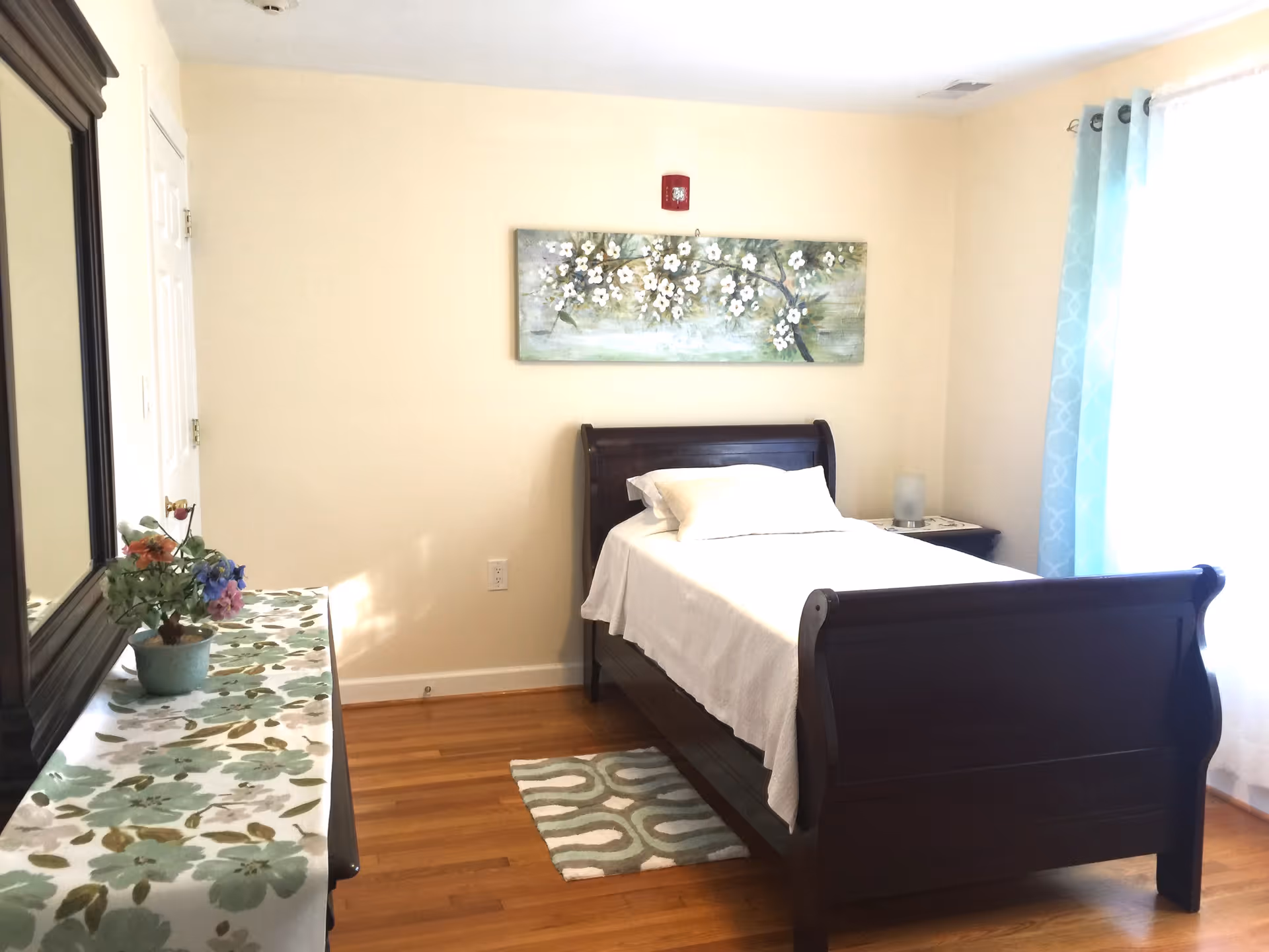 A bright bedroom with a dark wood sleigh bed made with white linens, a dresser with mirror and floral decor, wall art above the bed, and light blue curtains.