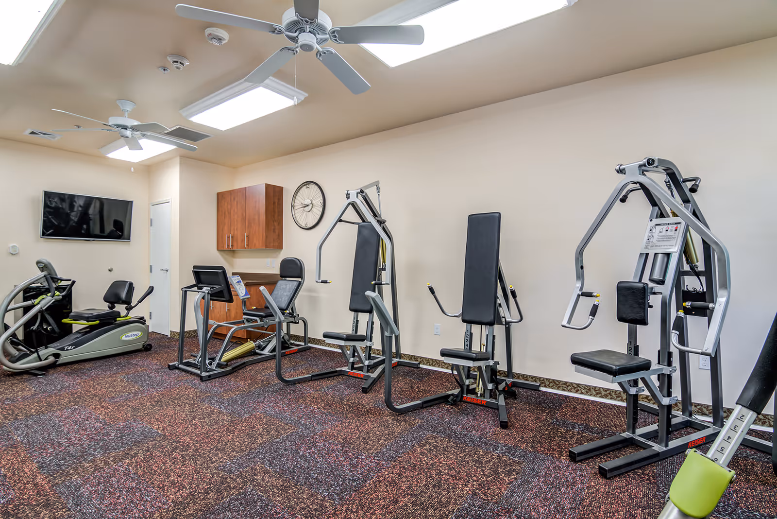 Small fitness room with several weight machines, cardio equipment, a wall-mounted TV and ceiling fans.