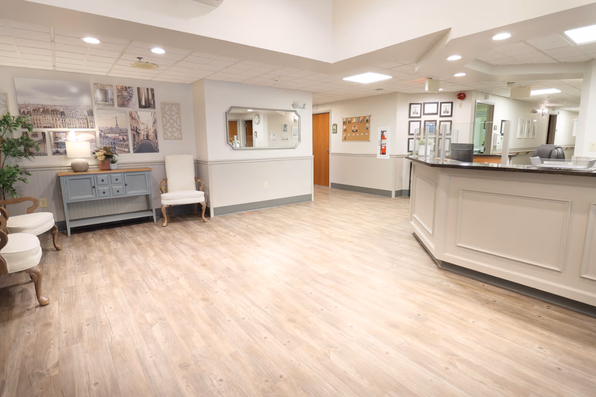 A spacious and well-lit reception area in a senior living facility with light wood flooring, a white reception desk on the right, several chairs along the left wall, a blue sideboard with a lamp and plant, and framed pictures on the walls.