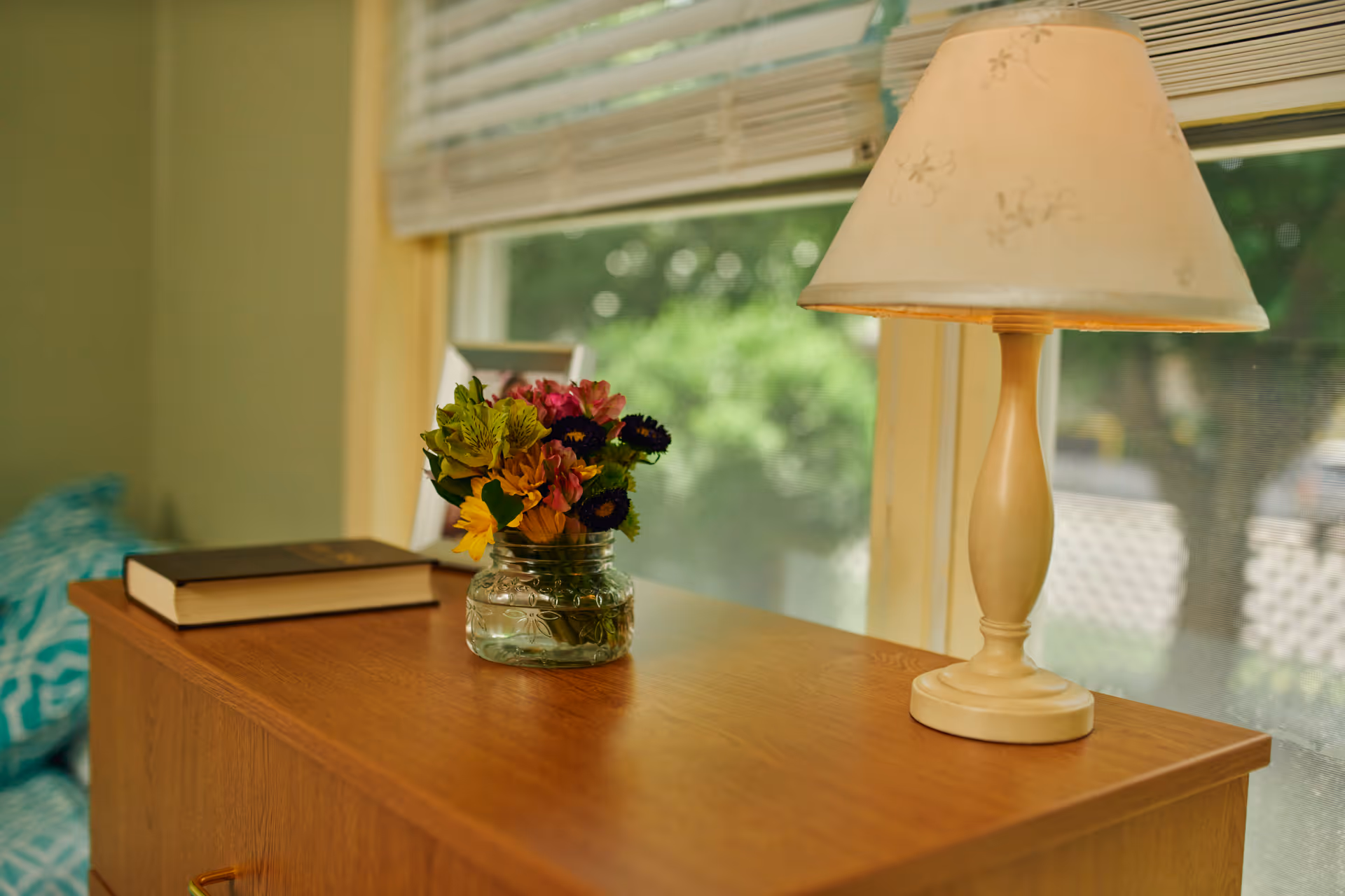 A wooden dresser with a small glass vase of colorful flowers, a closed book, and a beige table lamp with floral patterns, positioned in front of a window with white blinds partially open showing greenery outside.
