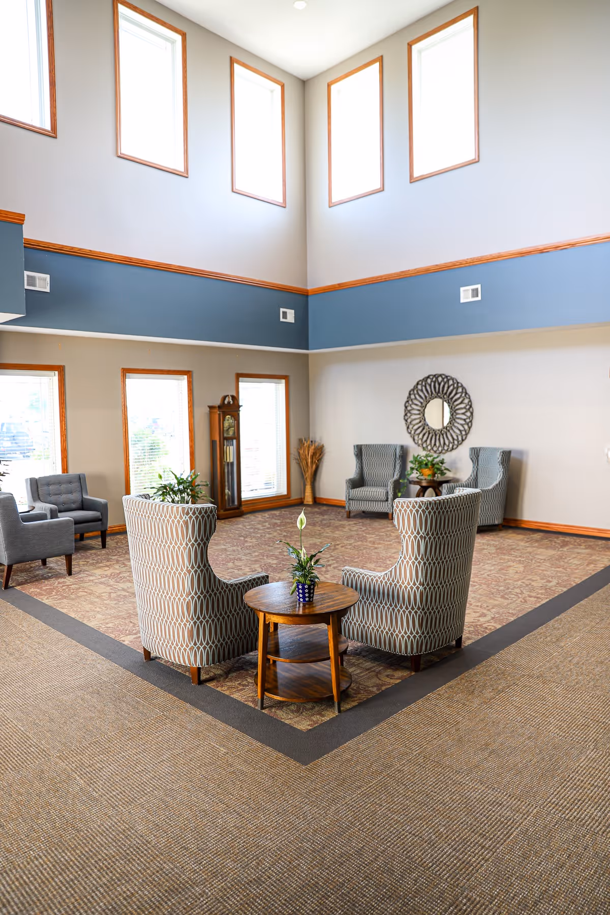 Spacious double-height common room with armchairs arranged around a small table, tall windows, and a decorative wall mirror.