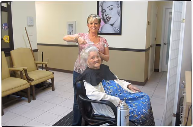 A woman is cutting the hair of an elderly woman seated in a chair inside a room with beige walls and tiled floor. There are two empty chairs against the wall and a framed black and white picture of Marilyn Monroe hanging on the wall behind them.