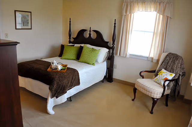 A cozy bedroom with a wooden bed frame, white bedding, and two green pillows. A brown blanket is folded at the foot of the bed with a tray holding a teapot and cup. There is a cushioned armchair with a floral pillow and a gray throw draped over it near a window with cream-colored curtains. A framed picture hangs on the wall and a wooden dresser is partially visible.