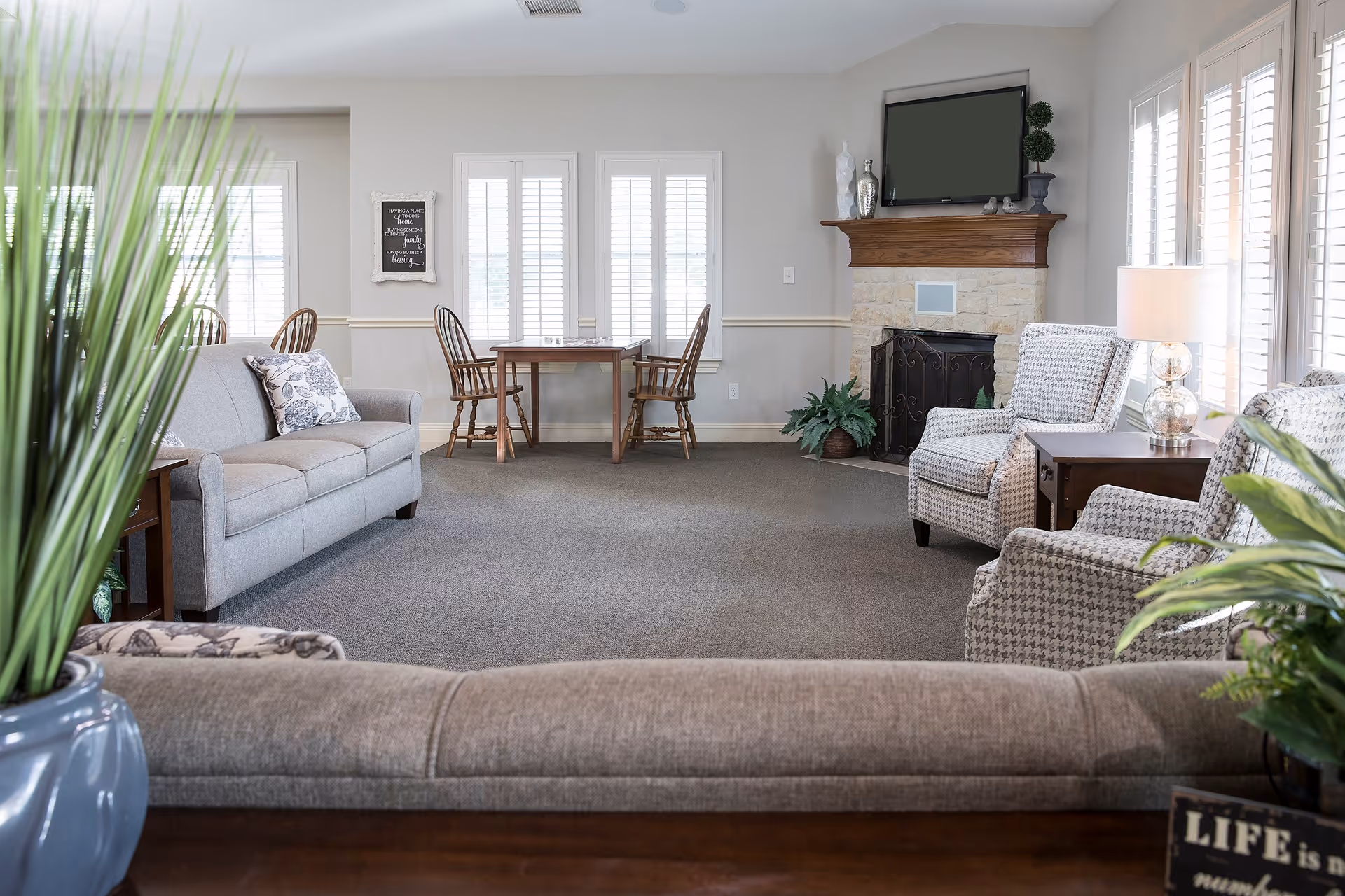 A bright and spacious living room with gray sofas and patterned armchairs arranged around a stone fireplace with a wooden mantle. A flat-screen TV is mounted above the fireplace. There is a wooden table with two chairs near the windows with white shutters, and several green plants add a touch of nature to the room.