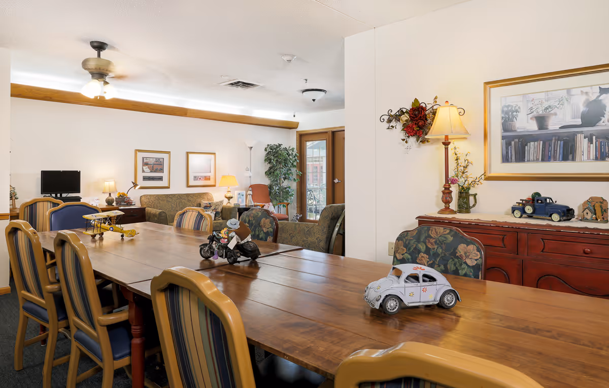 A cozy common area in an assisted living facility featuring a long wooden table with decorative model vehicles on it, surrounded by striped cushioned chairs. In the background, there are sofas, a small TV, lamps, framed pictures on the wall, a potted plant, and a door leading outside. A sideboard with a lamp, flowers, and more decorative items is visible on the right side under a framed picture of a cat sitting on a windowsill.