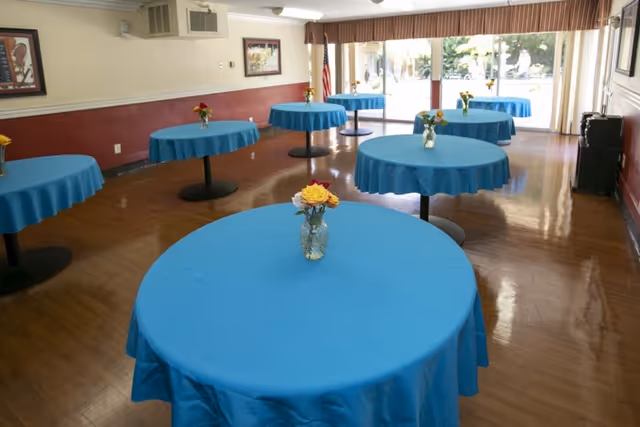 A dining room with several round tables covered with blue tablecloths, each table having a small vase with yellow and red flowers. The room has wooden flooring, framed pictures on the walls, and large windows with curtains letting in natural light.