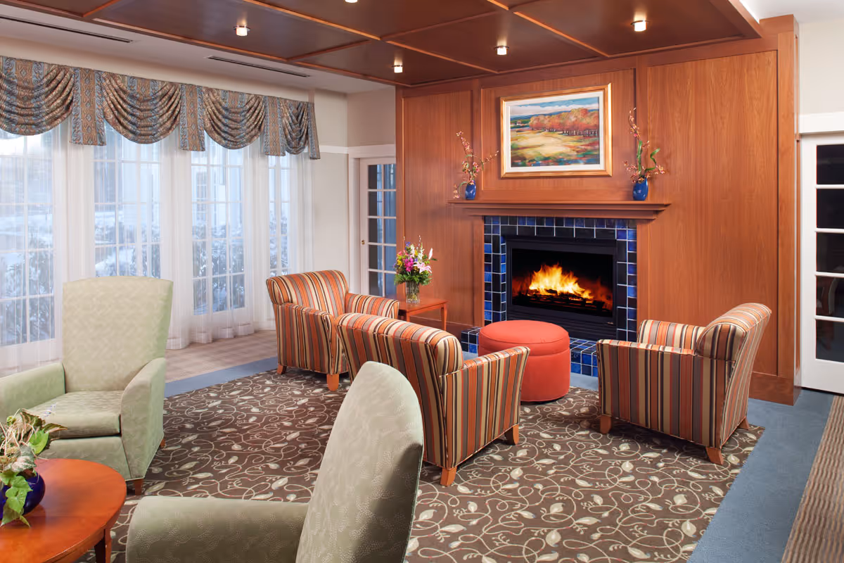 A cozy living room area with a lit fireplace surrounded by blue tiles and wooden paneling. Four striped armchairs and two green armchairs are arranged around a round red ottoman on a patterned carpet. Large windows with sheer curtains and decorative valances let in natural light. A colorful landscape painting hangs above the fireplace, and a small table with a flower vase is beside one of the armchairs.