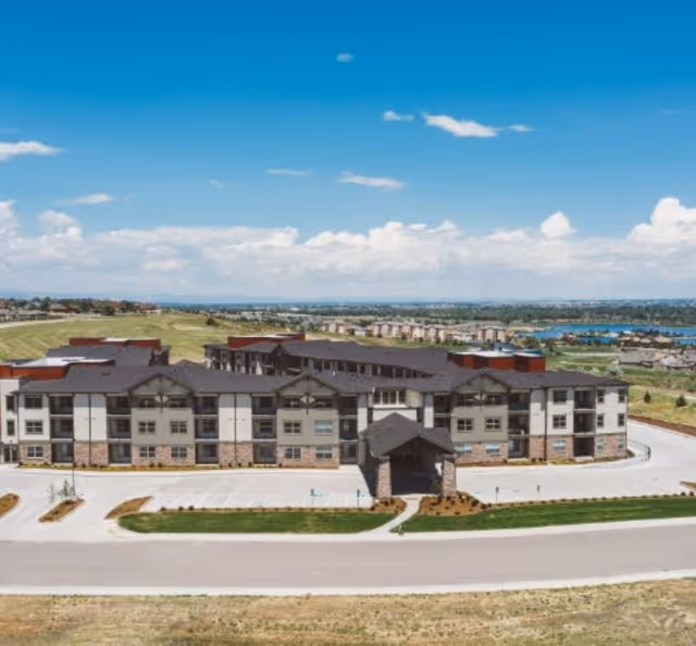 Front aerial view of a large multi-story senior living building with a covered entrance, surrounding parking and landscaped grounds under a blue sky.