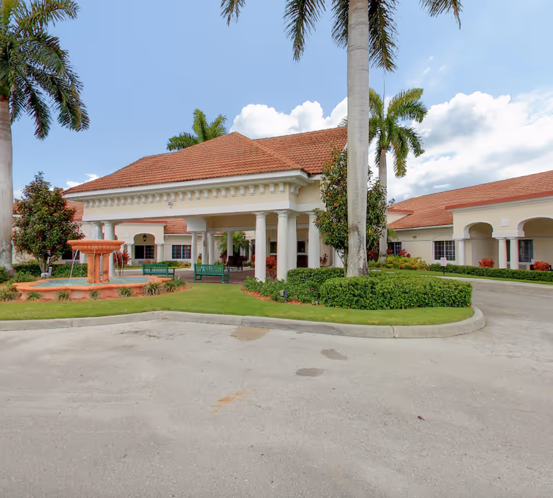 Entrance of a single-story Mediterranean-style senior living facility with a covered porte-cochere, palm trees, and a fountain in front.