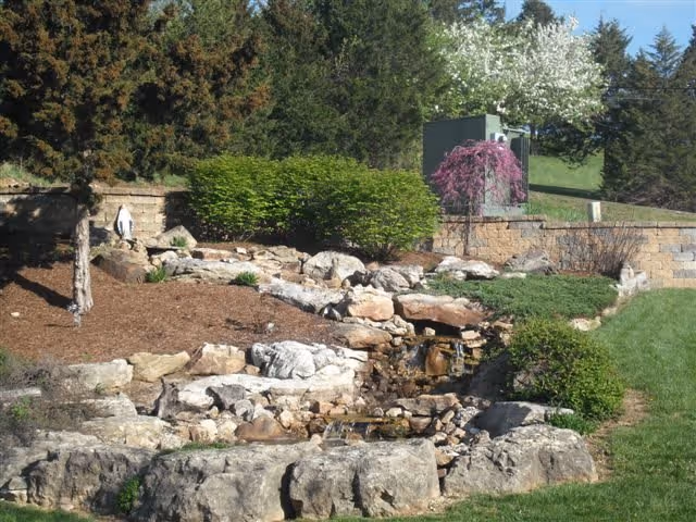 Landscaped outdoor area with rocks and a small cascading waterfall, shrubs, and trees.