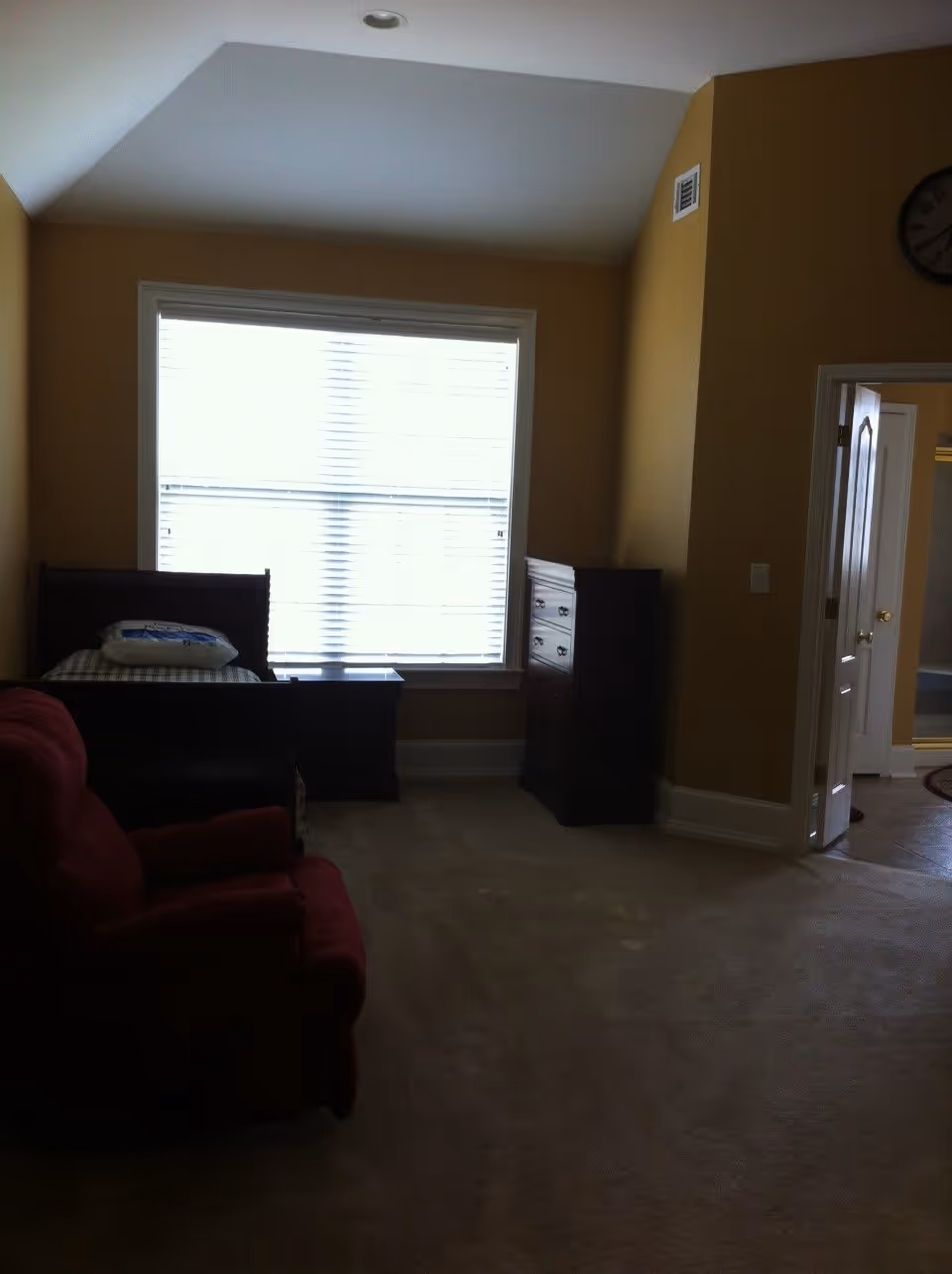 A bedroom with a single bed against the wall under a large window with blinds. There is a dark wooden nightstand next to the bed and a matching tall dresser against the adjacent wall. A red upholstered armchair is positioned in the foreground. The walls are painted a warm beige color and there is a doorway leading to a bathroom visible on the right side.