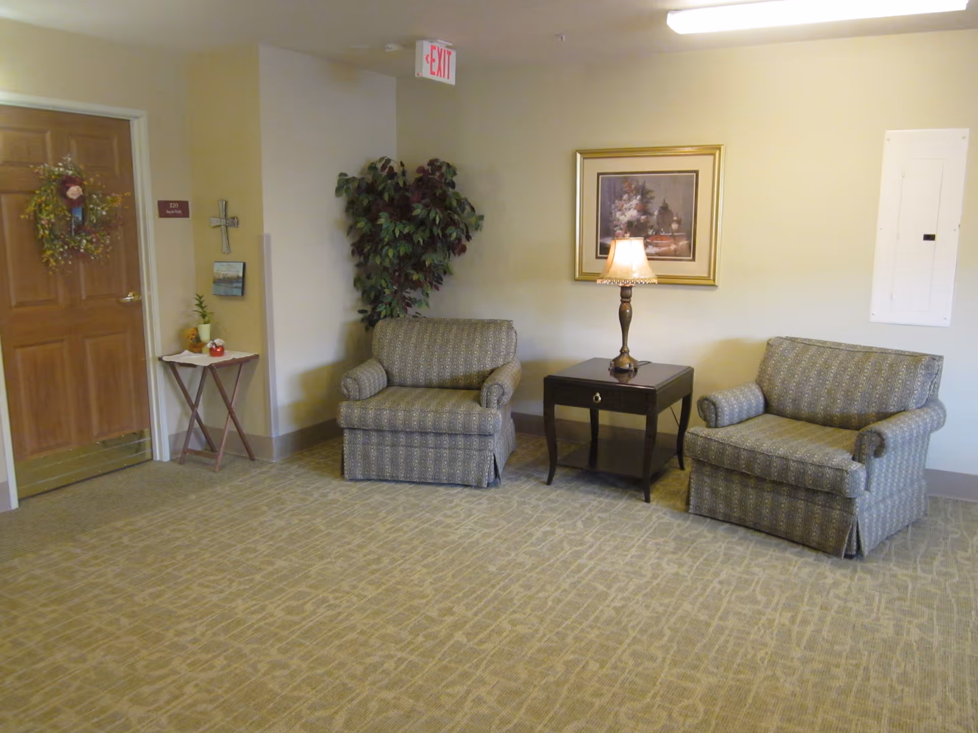 A cozy sitting area in a hallway with two patterned armchairs separated by a dark wooden side table with a lamp. A framed floral painting hangs on the wall above the table. To the left, there is a wooden door decorated with a wreath, a small table with plants and decorations, a cross, and a room number plaque. A large potted plant is placed in the corner near the chairs. The carpet has a subtle geometric pattern, and an exit sign is visible on the ceiling.
