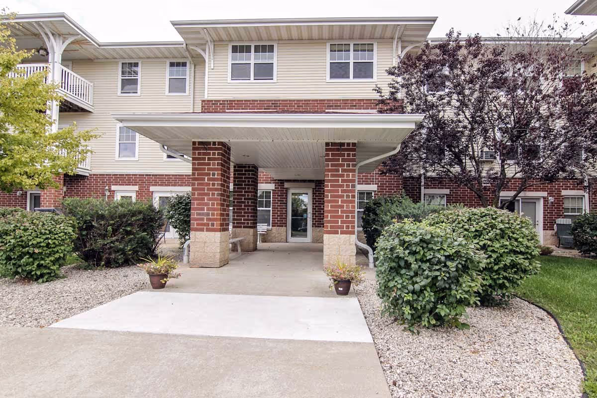 Entrance of a senior living facility with a covered walkway supported by brick pillars. The building has beige siding and red brick accents, with several windows visible. There are shrubs and small trees planted on either side of the walkway, along with two potted plants near the entrance.