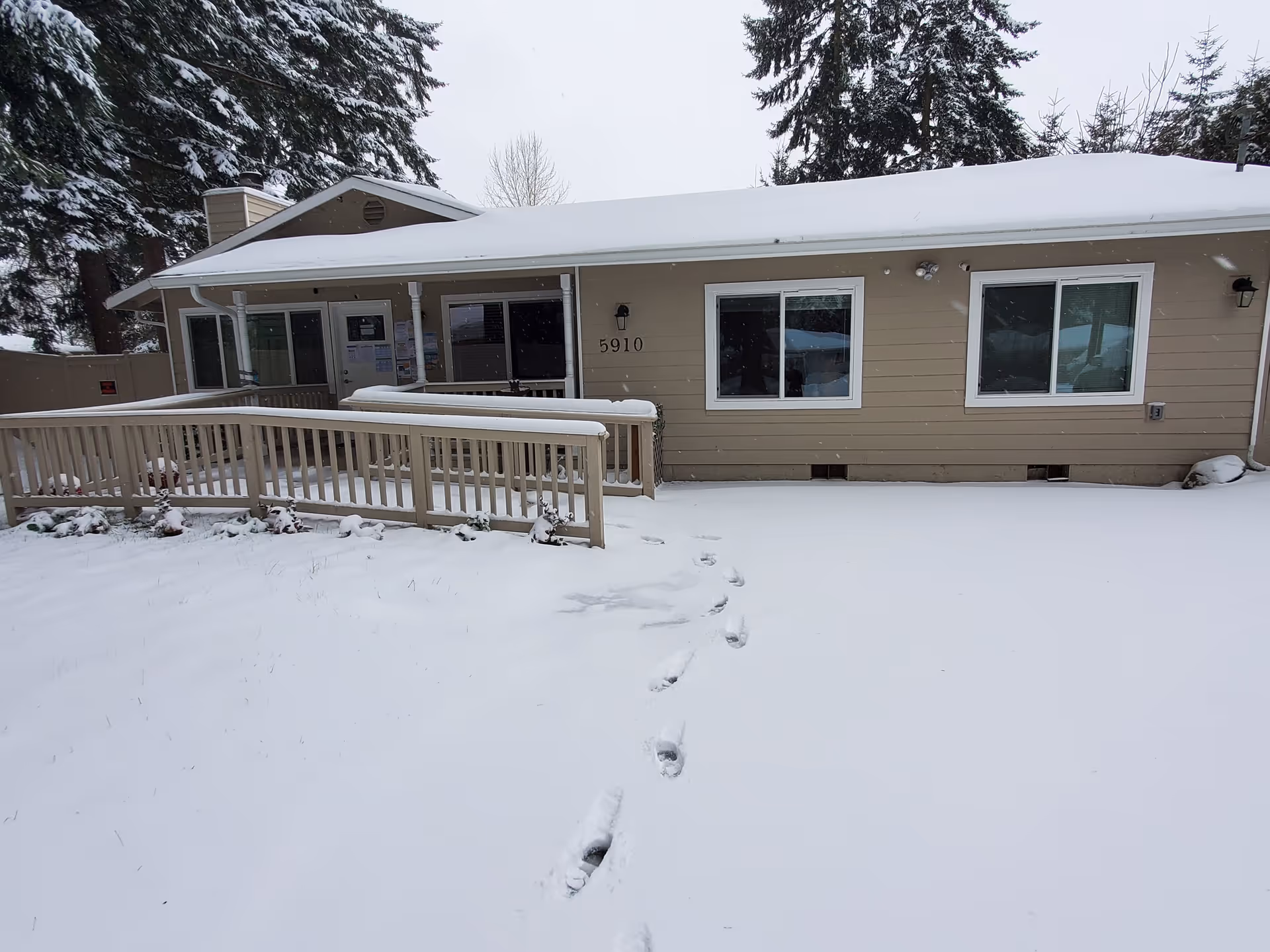 Single-story beige building with a snow-covered roof and ground. There is a wooden ramp leading to the entrance door, and footprints are visible in the snow leading to the building. The building number 5910 is displayed near the entrance. Tall evergreen trees are in the background.