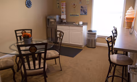 A small dining area with a round glass table surrounded by four chairs with cushioned seats. There is a countertop with three high chairs on the right side, and a soft-serve ice cream cone decoration on the counter. Against the far wall, there is a water dispenser, a cabinet with a coffee maker, and a trash can near a window with blinds. The walls have a few posters and a clock.