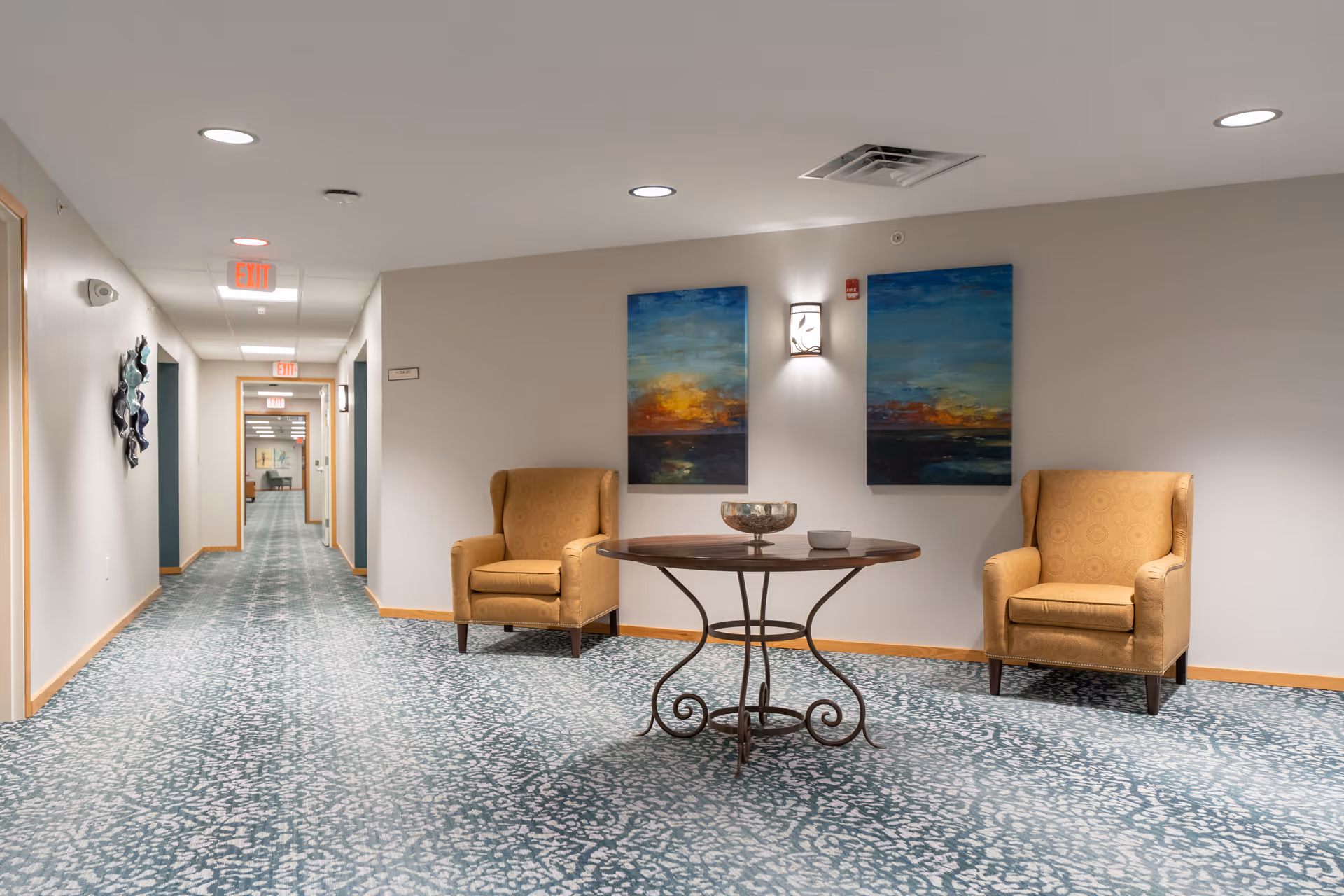 Carpeted corridor seating area with two upholstered armchairs, a round table, and landscape paintings on the wall.