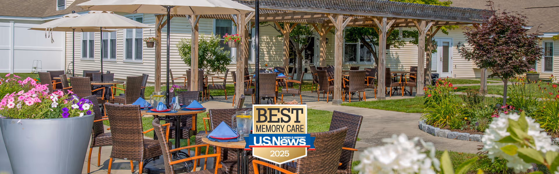 Outdoor seating area at The Arbors at Shelburne with multiple round tables and wicker chairs under large umbrellas and a wooden pergola. There are colorful flowers in planters and landscaped greenery surrounding the paved patio space. A sign in the center reads 'Best Memory Care U.S. News 2025'.