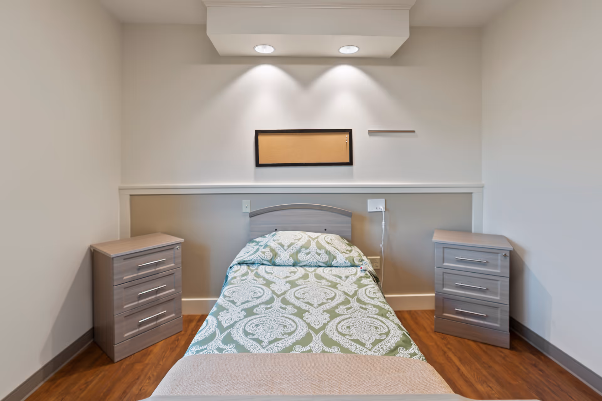 A simple, clean bedroom with a single bed centered against the wall. The bed has a green and white patterned bedspread and a beige blanket at the foot. On either side of the bed are matching gray nightstands with three drawers each. Above the bed is a small cork bulletin board mounted on the wall, illuminated by two recessed ceiling lights. The room has wood flooring and neutral-colored walls.