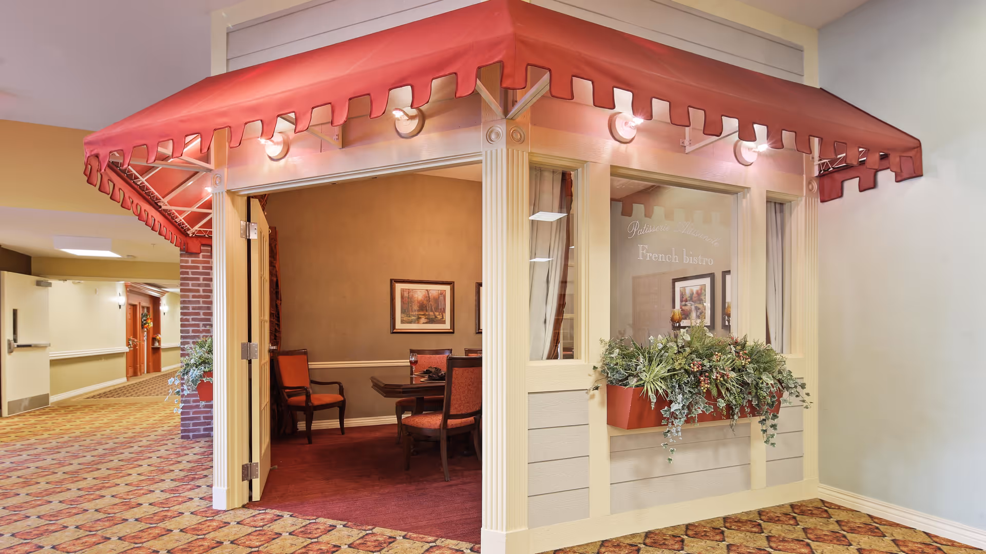 Indoor hallway with patterned carpet leading to a small room styled like a French bistro with a red awning, window box with greenery, and a table with chairs inside.