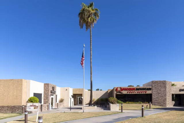 Front exterior of the Phoenix Mountain facility showing the building entrance, an American flag on a pole, and a tall palm tree under a clear blue sky.