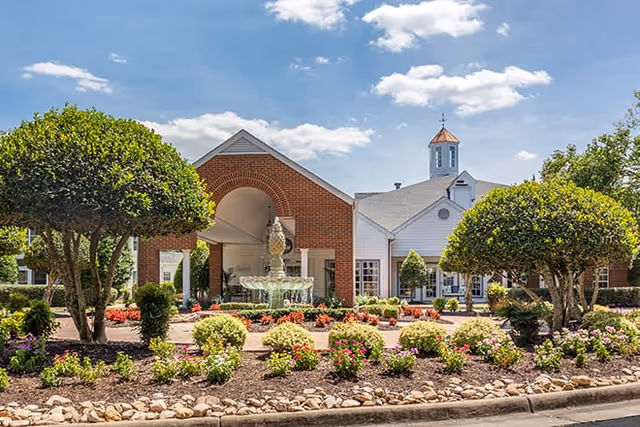 Brick and white senior living facility entrance with a central fountain, landscaped flower beds, and trimmed trees under a blue sky.