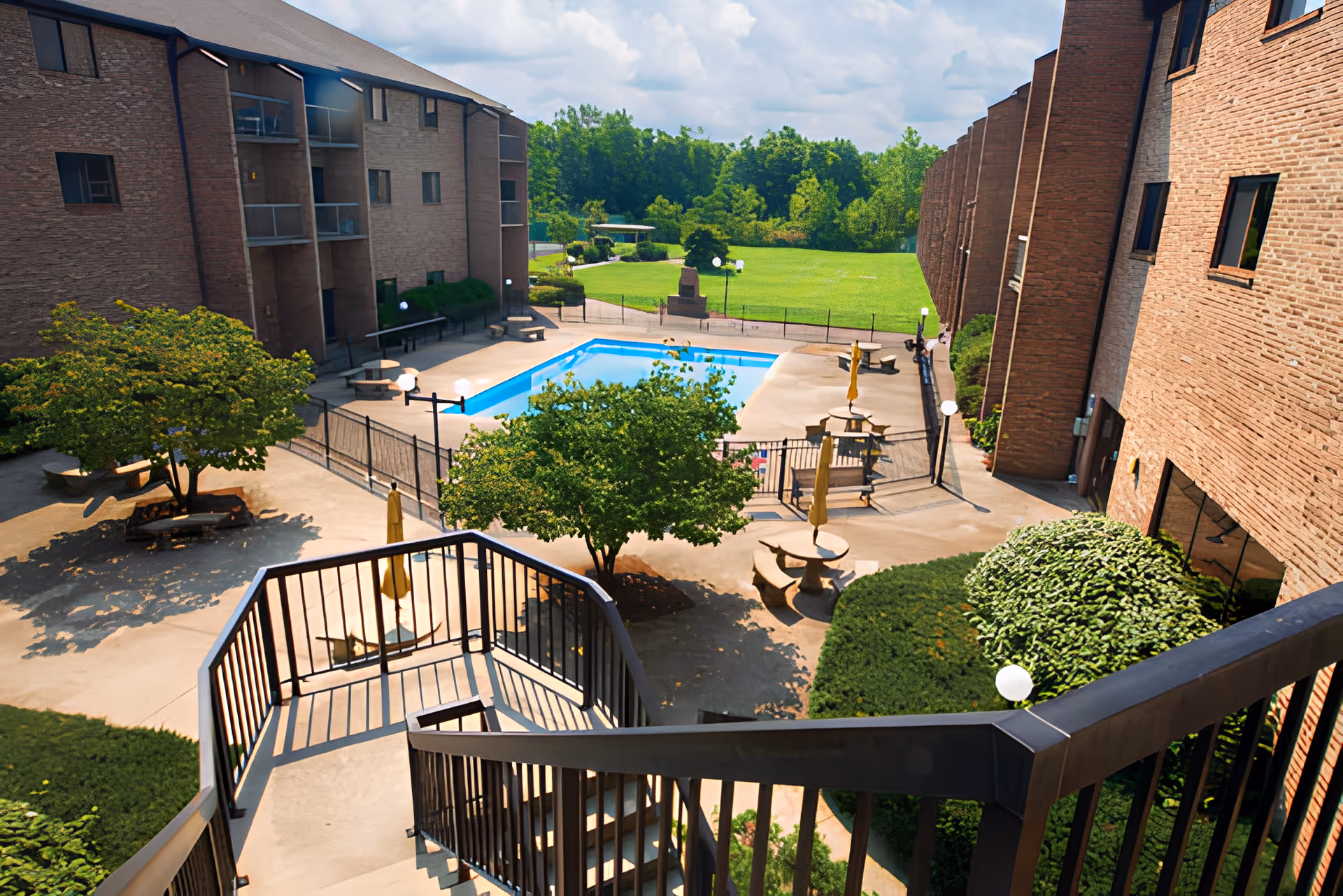 View from a staircase overlooking an outdoor courtyard area with a swimming pool, surrounded by brick buildings. The courtyard has several trees, benches, tables with umbrellas, and well-maintained greenery. In the background, there is a large grassy area with trees and a partly cloudy sky.