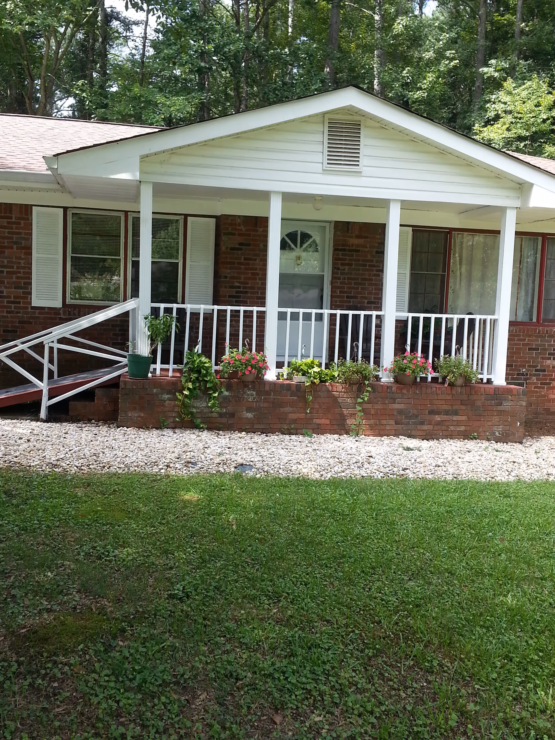 Front porch of a brick house with white railings and columns, a white door with a window, several potted plants with flowers on the brick ledge, a wheelchair ramp on the left side, and green grass in the foreground with trees in the background.
