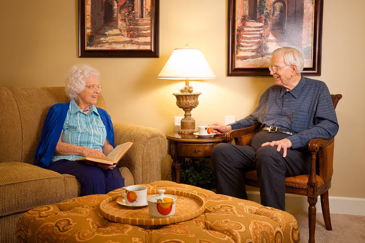 An elderly woman sitting on a beige couch holding an open book, smiling and talking to an elderly man seated in a wooden armchair. Between them is a small wooden side table with a lamp and two cups. The room has warm lighting and two framed paintings on the wall behind them.