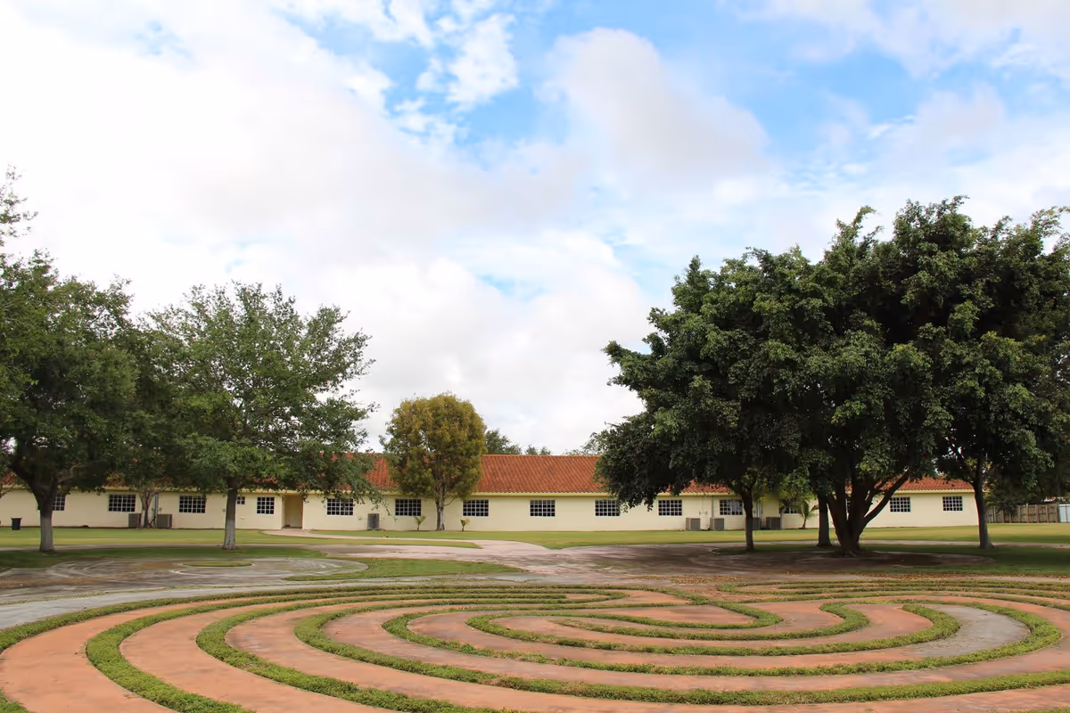 Outdoor view of a garden area with a circular labyrinth pattern made of grass and concrete paths, surrounded by several large green trees and a single-story building with a red-tiled roof in the background under a partly cloudy sky.