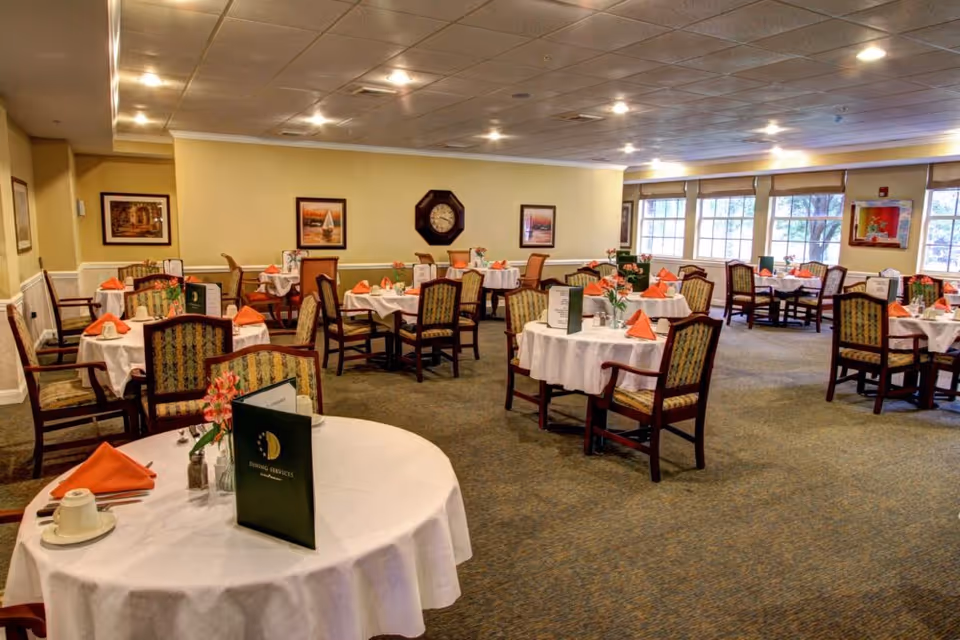 Bright dining room with round tables set with white tablecloths, menus, orange napkins, and chairs.