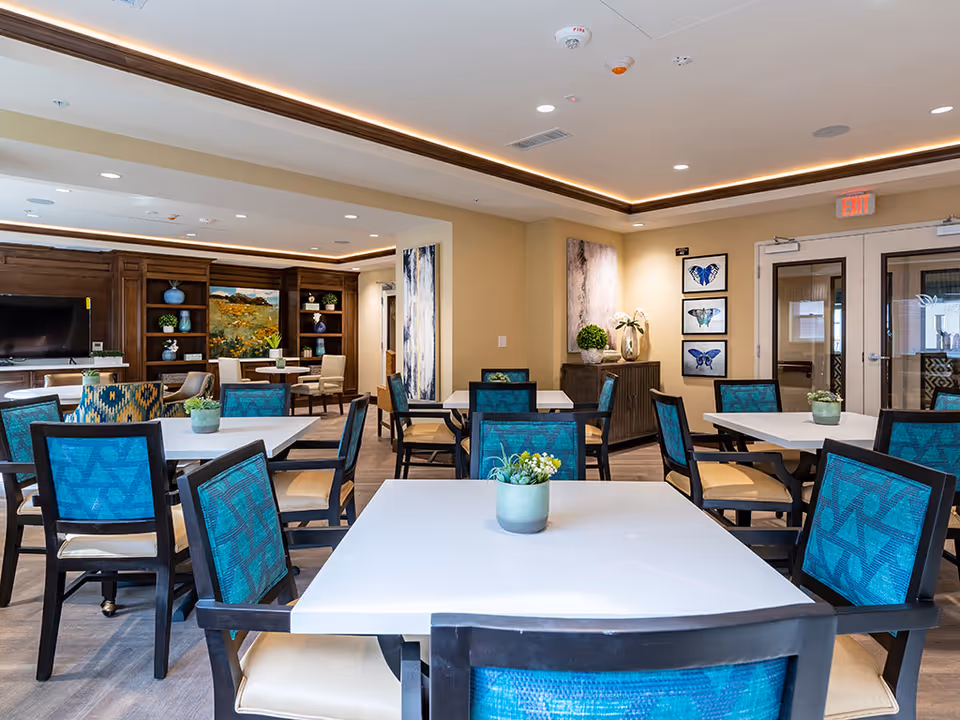Communal dining room with white square tables, teal-upholstered chairs, potted plants, and a wood-paneled seating area with TV and artwork.