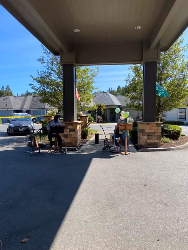 Two musicians playing instruments under a covered entrance area outside a residential building with trees and parked cars in the background on a sunny day.