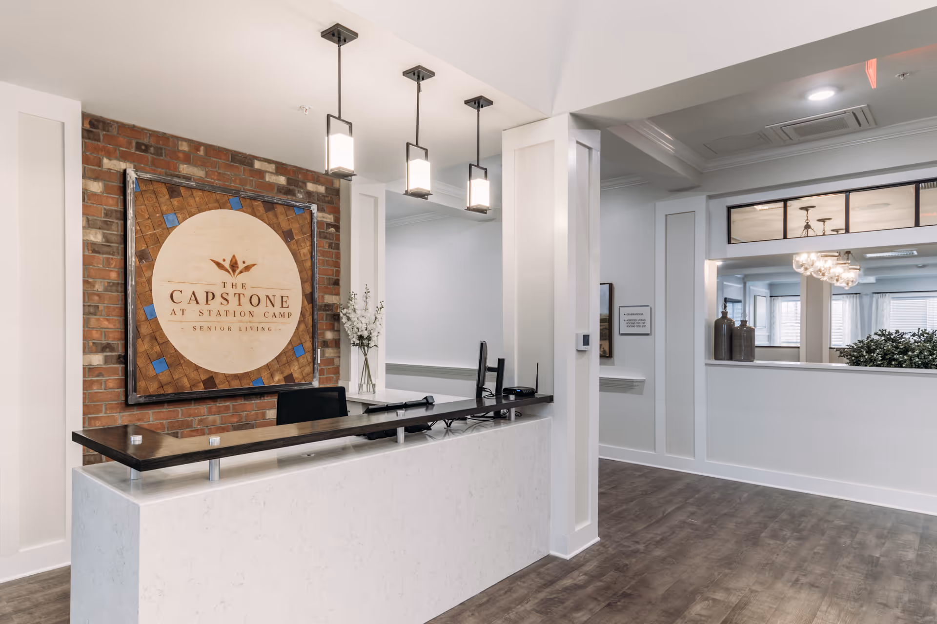 Reception area of The Capstone at Station Camp senior living facility featuring a modern white marble desk with a dark wood countertop, three pendant lights hanging from the ceiling, a brick accent wall with a large wooden sign displaying the facility's name, and a hallway with decorative vases and a large mirror.