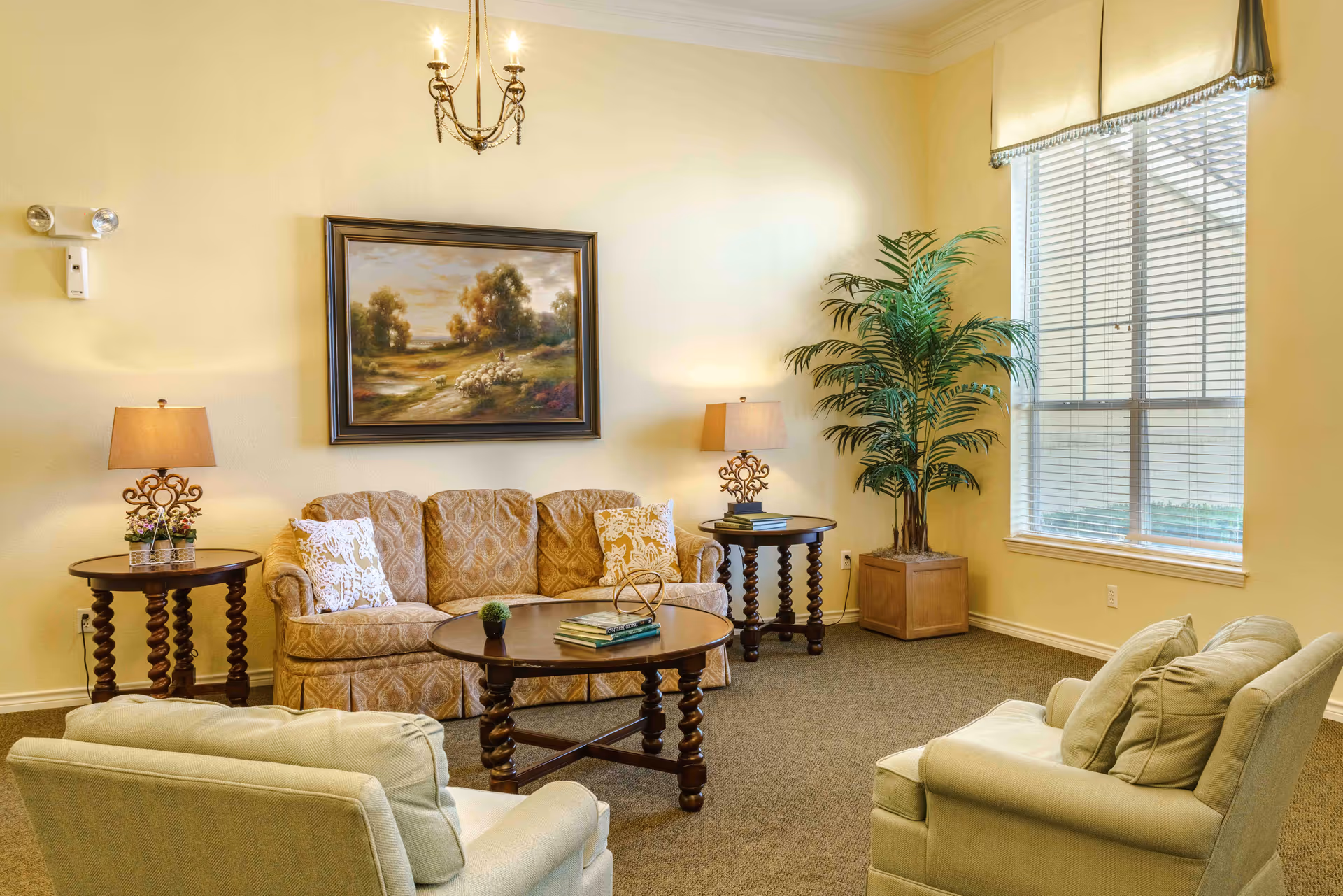 A cozy living room with a patterned sofa, two cushioned armchairs, a round wooden coffee table with books and a small plant, two matching side tables with lamps, a large potted plant near a window with blinds, and a framed painting of a pastoral scene on the wall.