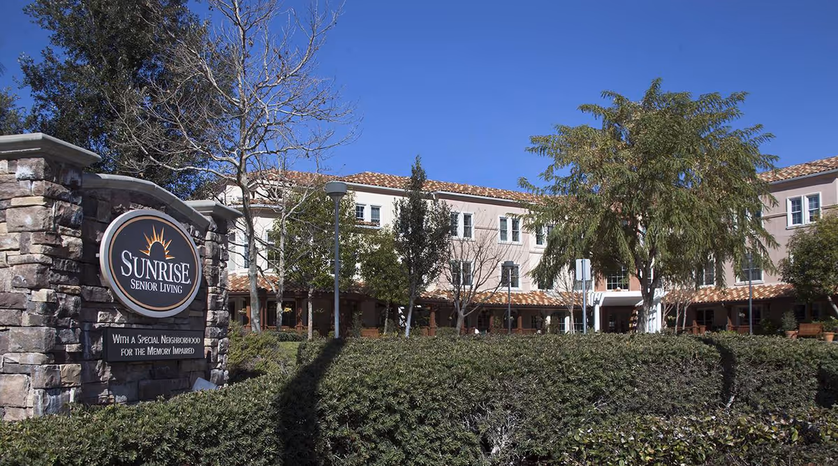 Front exterior of the Sunrise Senior Living facility showing the building, stone entrance sign, and landscaping under a clear sky.