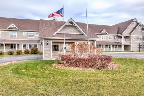 Exterior view of a large senior living facility building with beige siding and a brown roof. There is a covered entrance with two flagpoles in front, one flying the American flag. The foreground features a circular landscaped area with bushes and grass.