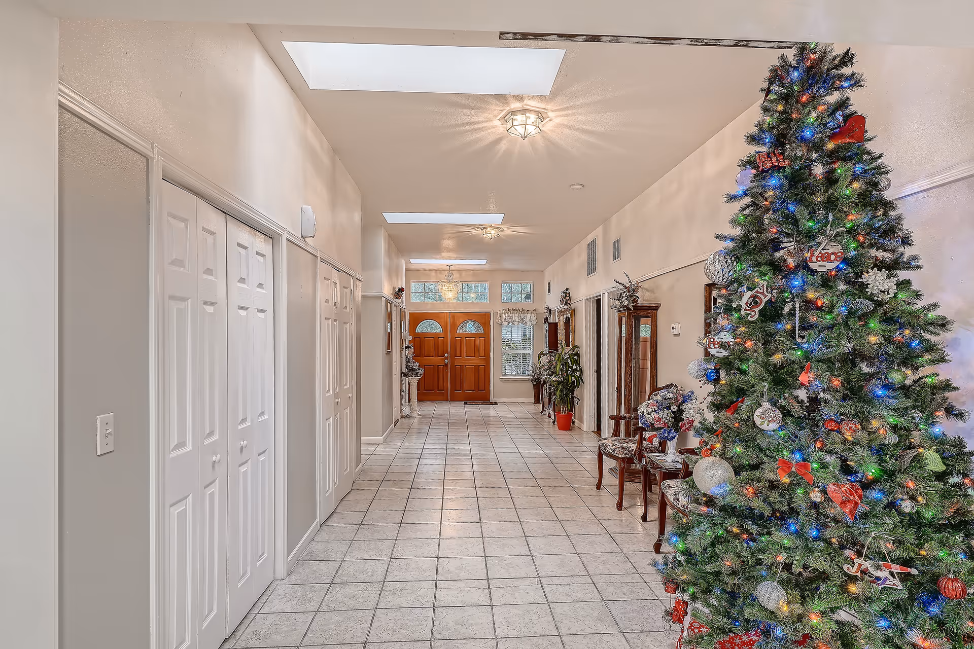 Tiled foyer hallway decorated with a large Christmas tree, chairs, and plants leading to double wooden front doors.
