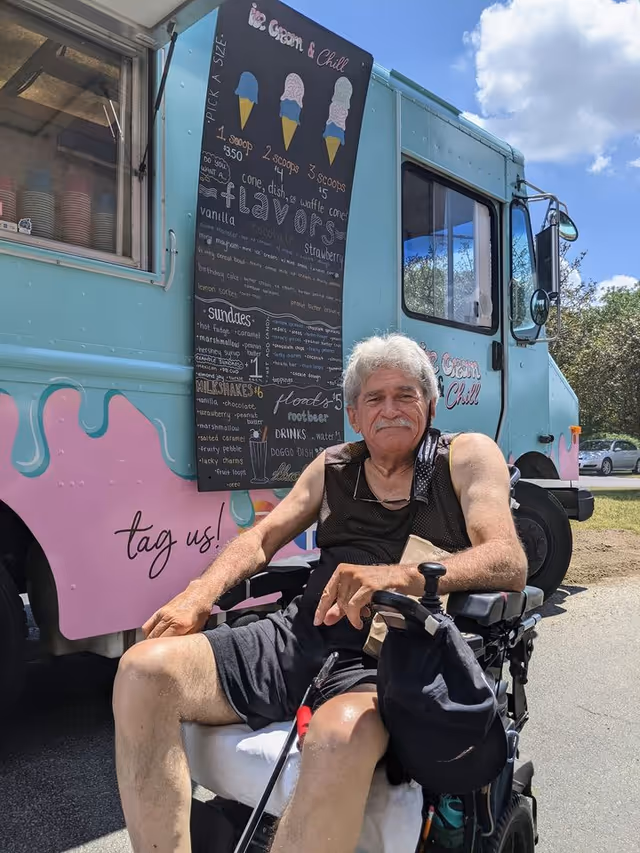 An elderly man in a wheelchair is sitting outdoors in front of a pastel blue and pink ice cream truck with a menu board displaying ice cream flavors and prices. The man is wearing a black sleeveless shirt and shorts, and there are trees and a partly cloudy sky in the background.