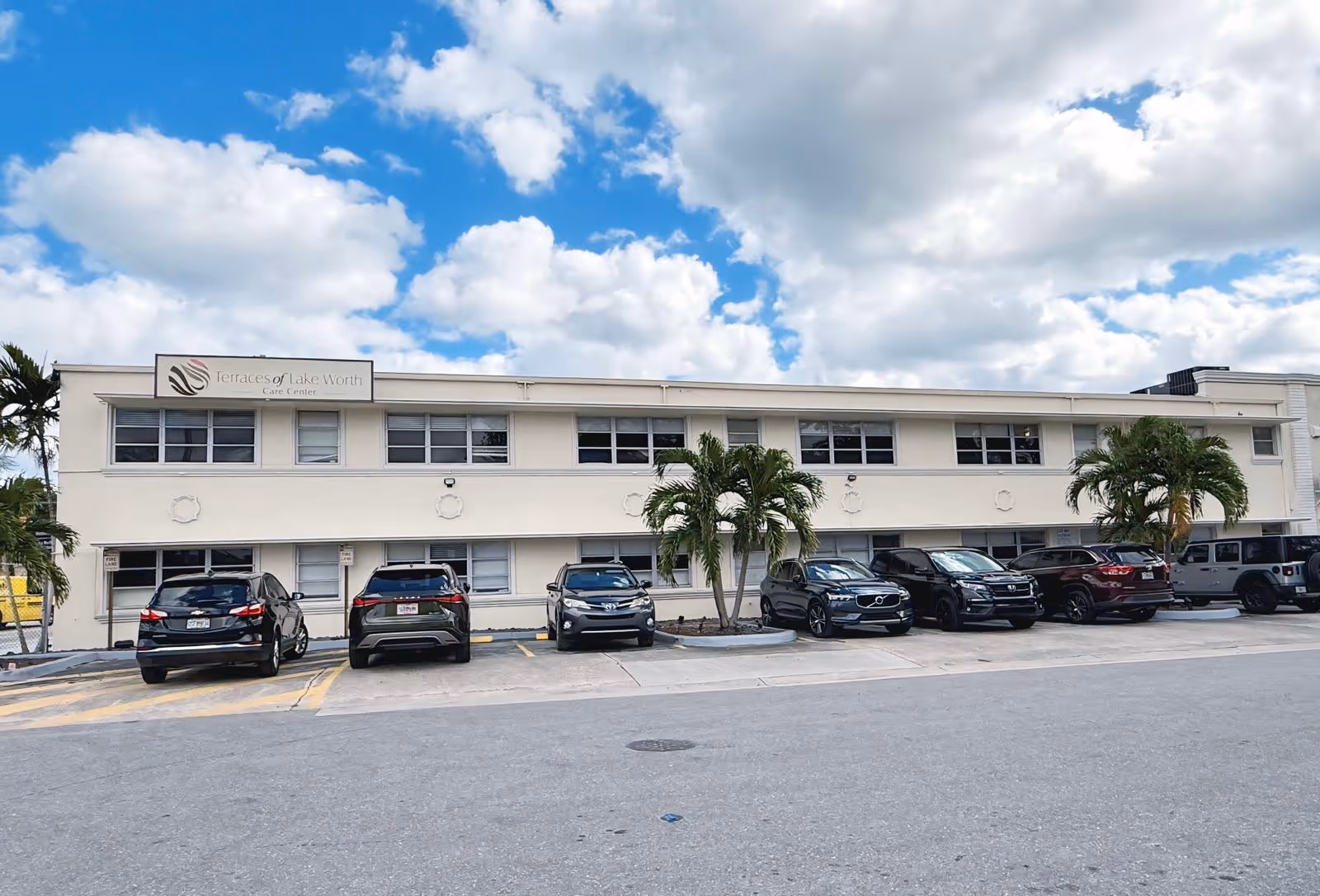 Exterior view of a two-story beige building with multiple windows and a sign that reads 'Terraces of Lake Worth Care Center'. Several cars are parked in front of the building, and there are palm trees in the parking area. The sky is partly cloudy with blue patches.