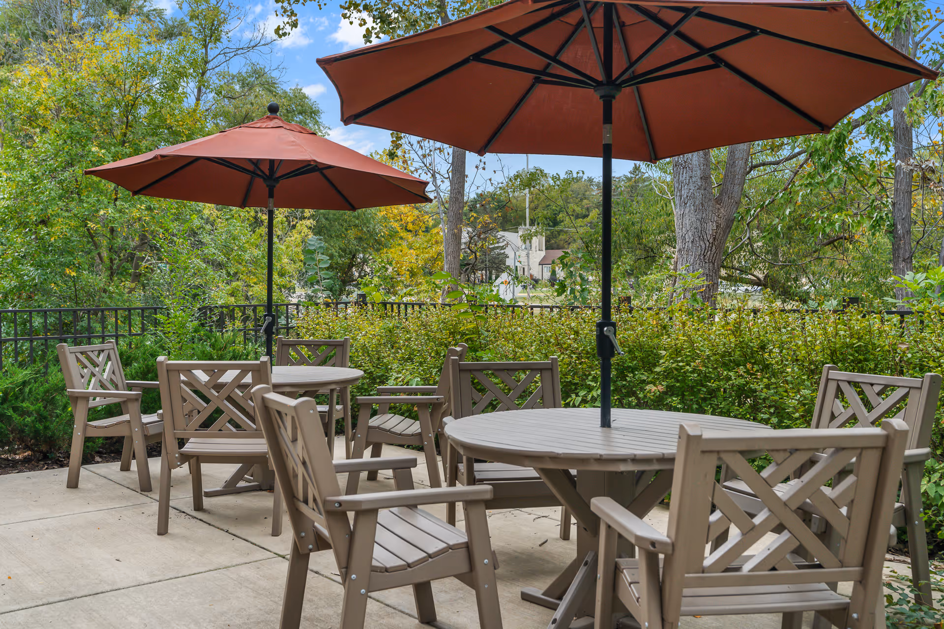 Outdoor patio with round tables, wooden chairs, and red umbrellas surrounded by trees and shrubbery.