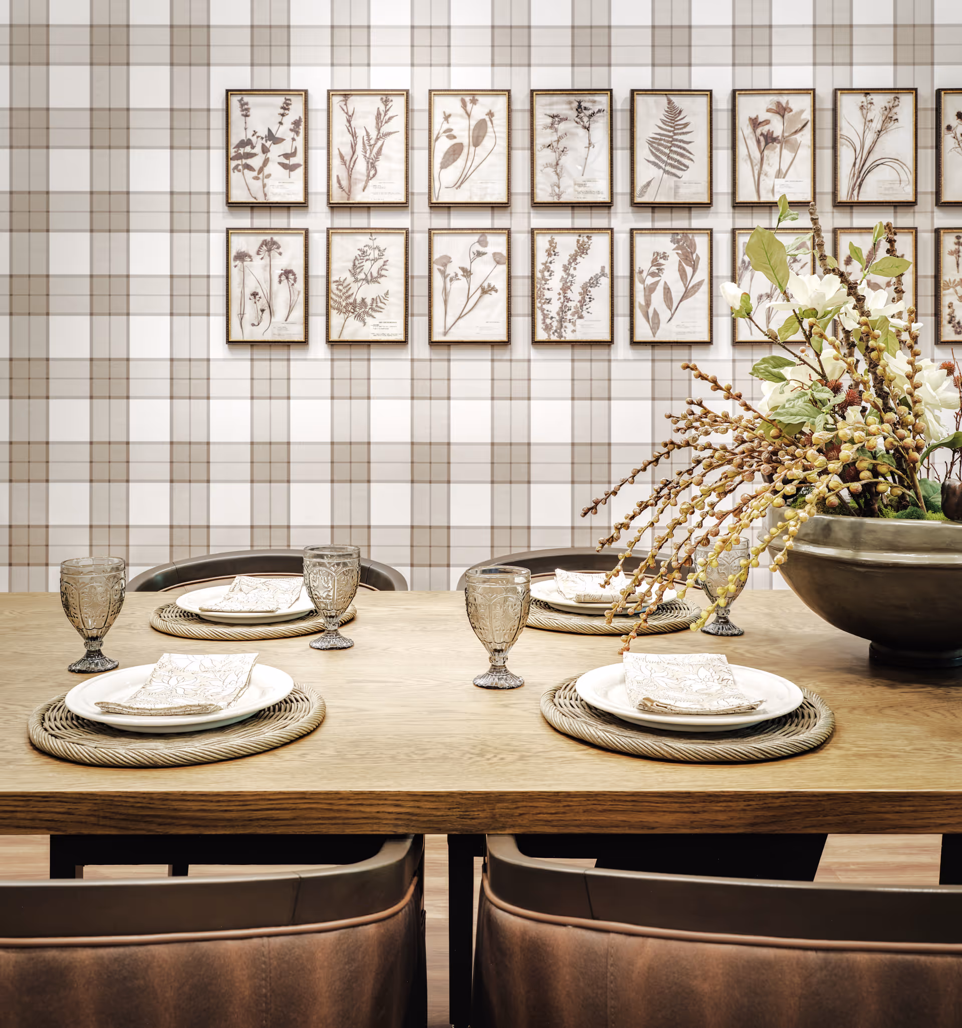 A dining table set for four with plates, napkins, and glass goblets on woven placemats. A large floral arrangement in a bowl is on the right side of the table. The background features a plaid patterned wall with framed botanical prints arranged in two rows.