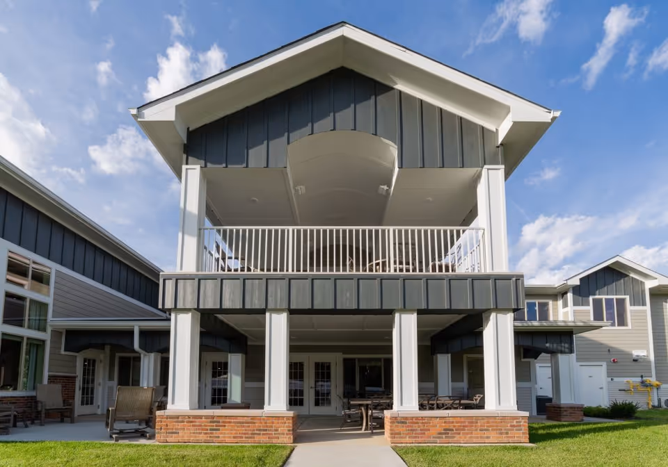 Front exterior of a two-story senior living facility showing a covered balcony, white columns, brick bases and patio seating.