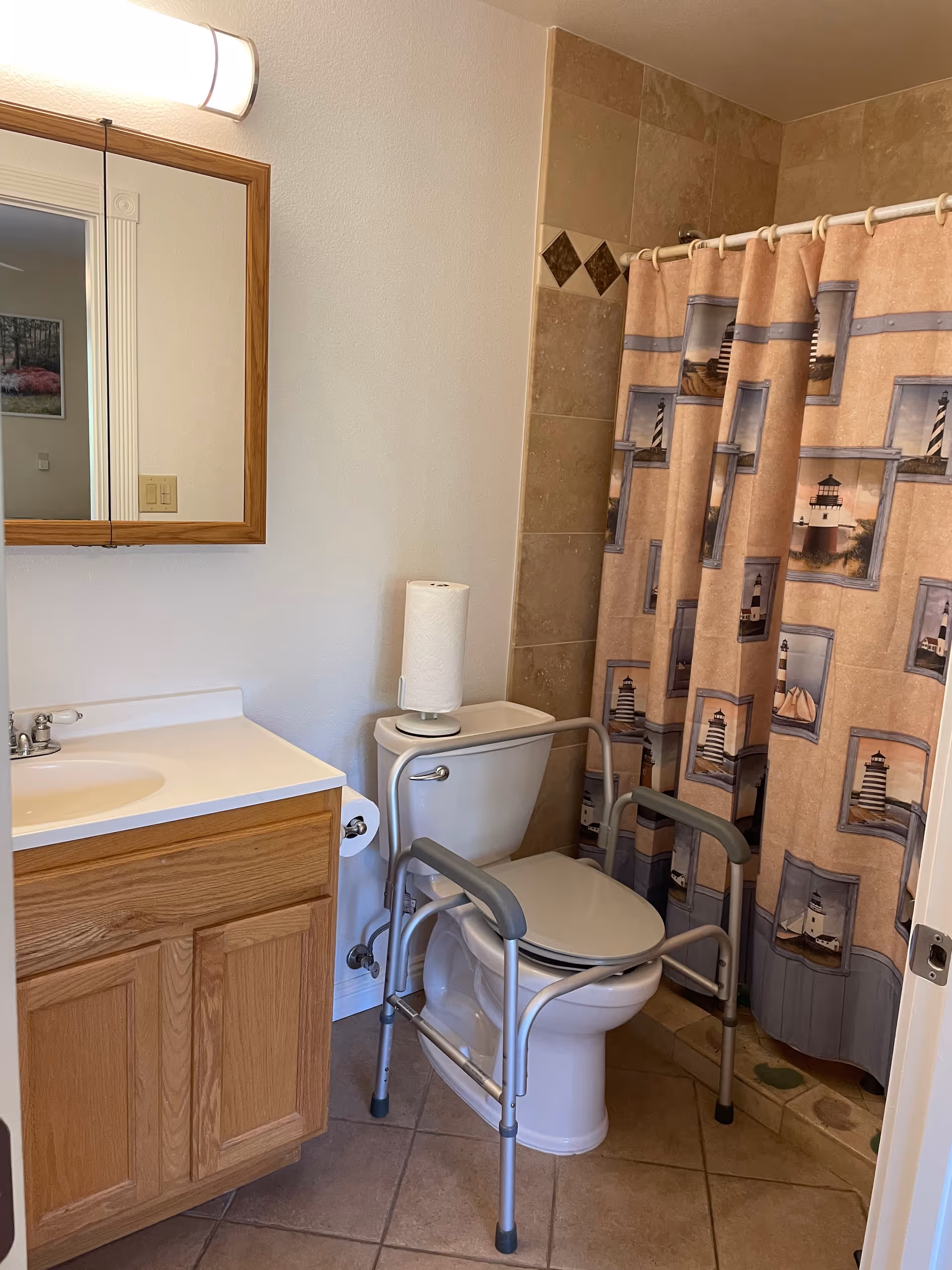 A bathroom with a wooden vanity and sink on the left, a toilet with a safety frame around it, and a shower with a curtain featuring lighthouse images. There is a roll of paper towels on top of the toilet tank and a mirror above the sink.