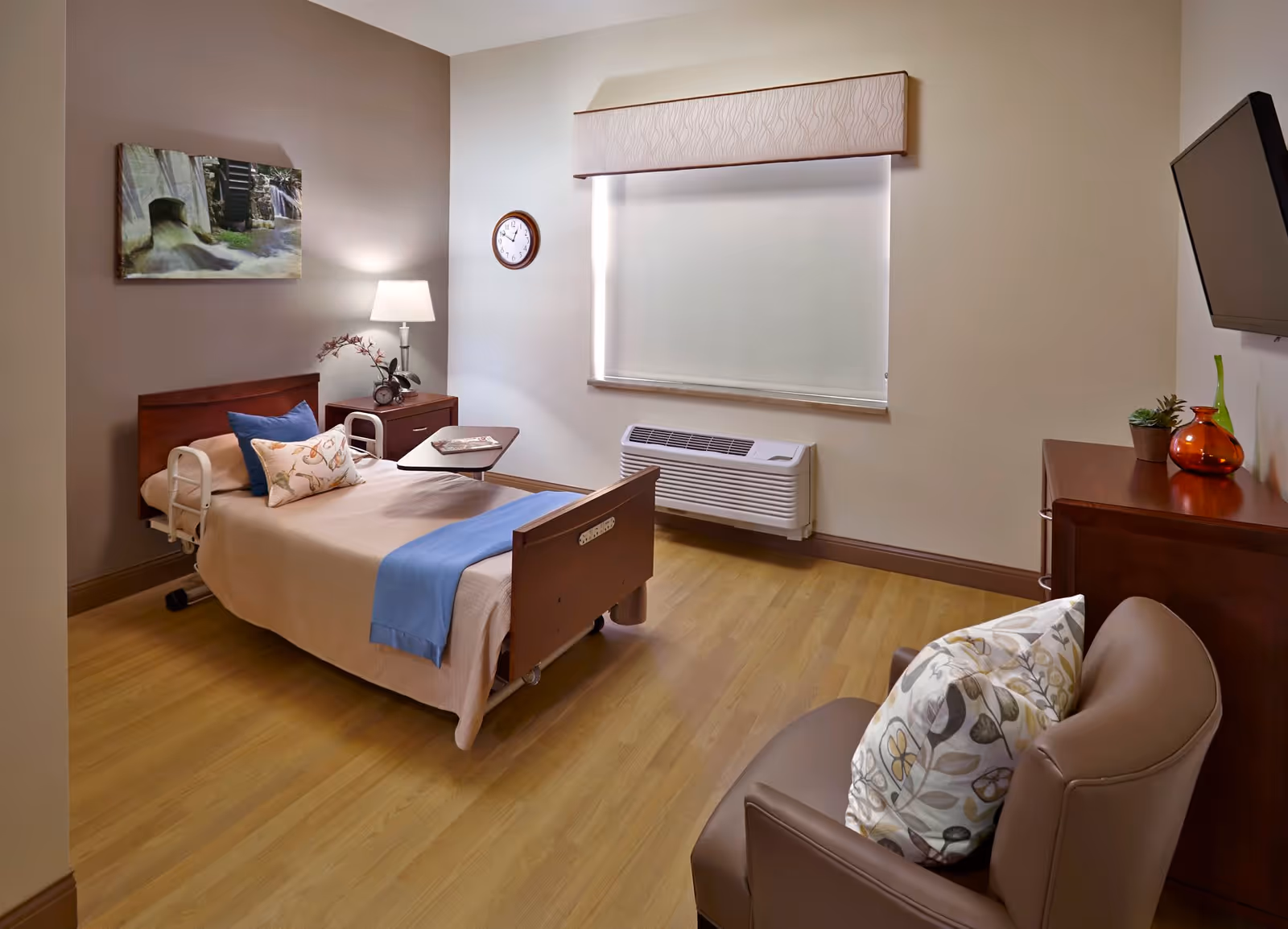 A clean and well-lit bedroom in a senior living facility featuring a single hospital-style bed with beige bedding and blue accents, a bedside table with a lamp and decorative items, a wall clock, a window with a closed blind, a mounted flat-screen TV, a wooden dresser with decorative vases, and a comfortable armchair with a patterned pillow.