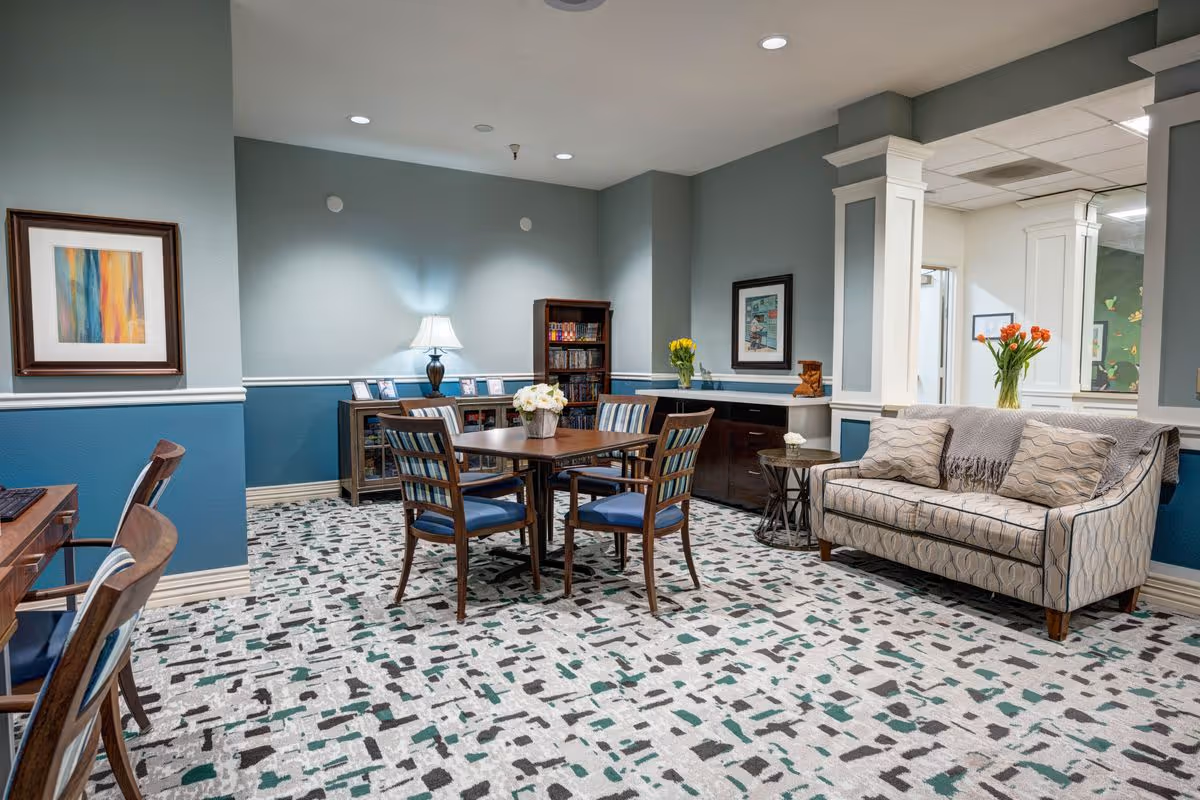 A cozy common area in a senior living facility featuring a patterned carpet, a wooden table with four chairs, a beige patterned sofa with two pillows and a throw blanket, a side table with a flower vase, framed artwork on the walls, a lamp on a cabinet, and a bookshelf filled with books.