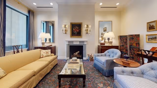 A cozy living room with a yellow sofa on the left and two light blue armchairs on the right. A glass coffee table with books is centered on a patterned rug. The room features a white fireplace with decorative sconces and a floral painting above it. There are two wooden side tables with lamps and mirrors on either side of the fireplace. Large windows with beige curtains allow natural light to enter the room.