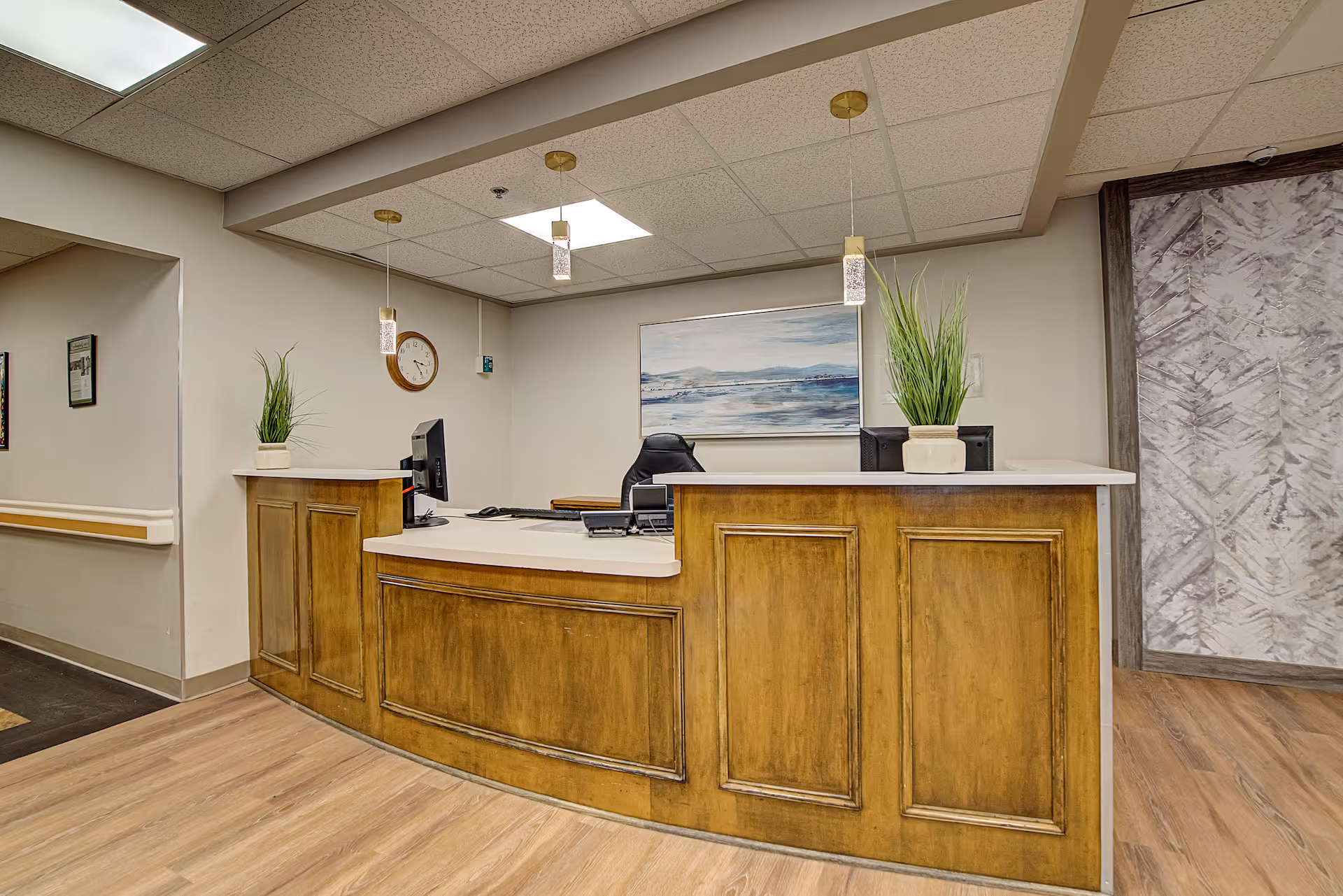 Reception desk area with a wooden counter, two computer monitors, two potted plants, a wall clock, and a landscape painting on the wall behind. The floor is wood laminate and the ceiling has recessed lighting and hanging pendant lights.